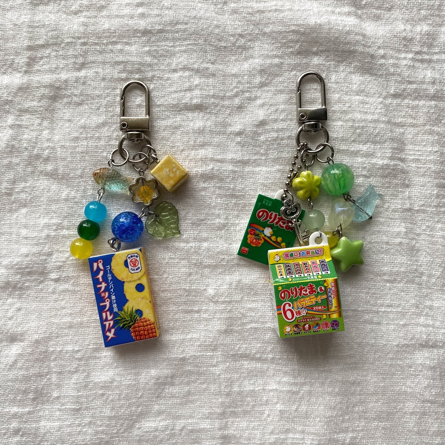 two novelty keychains are displayed on a textured white surface. the keychain on the left features a miniature box of pineapple candy, various colored beads, and other small charms. the one on the right has a miniature box of noritama and other charms and beads.