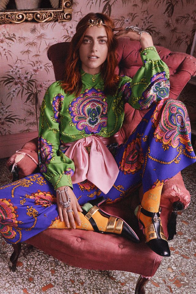 a fashion photograph shows a female model seated in a pink armchair. she is wearing a vibrant green and purple paisley patterned outfit consisting of a longsleeved blouse and widelegged pants. a pink sash is tied around her waist. she also sports gold shoes and a gold headpiece. the background is a floral wallpaper. the overall style is reminiscent of 70s fashion.