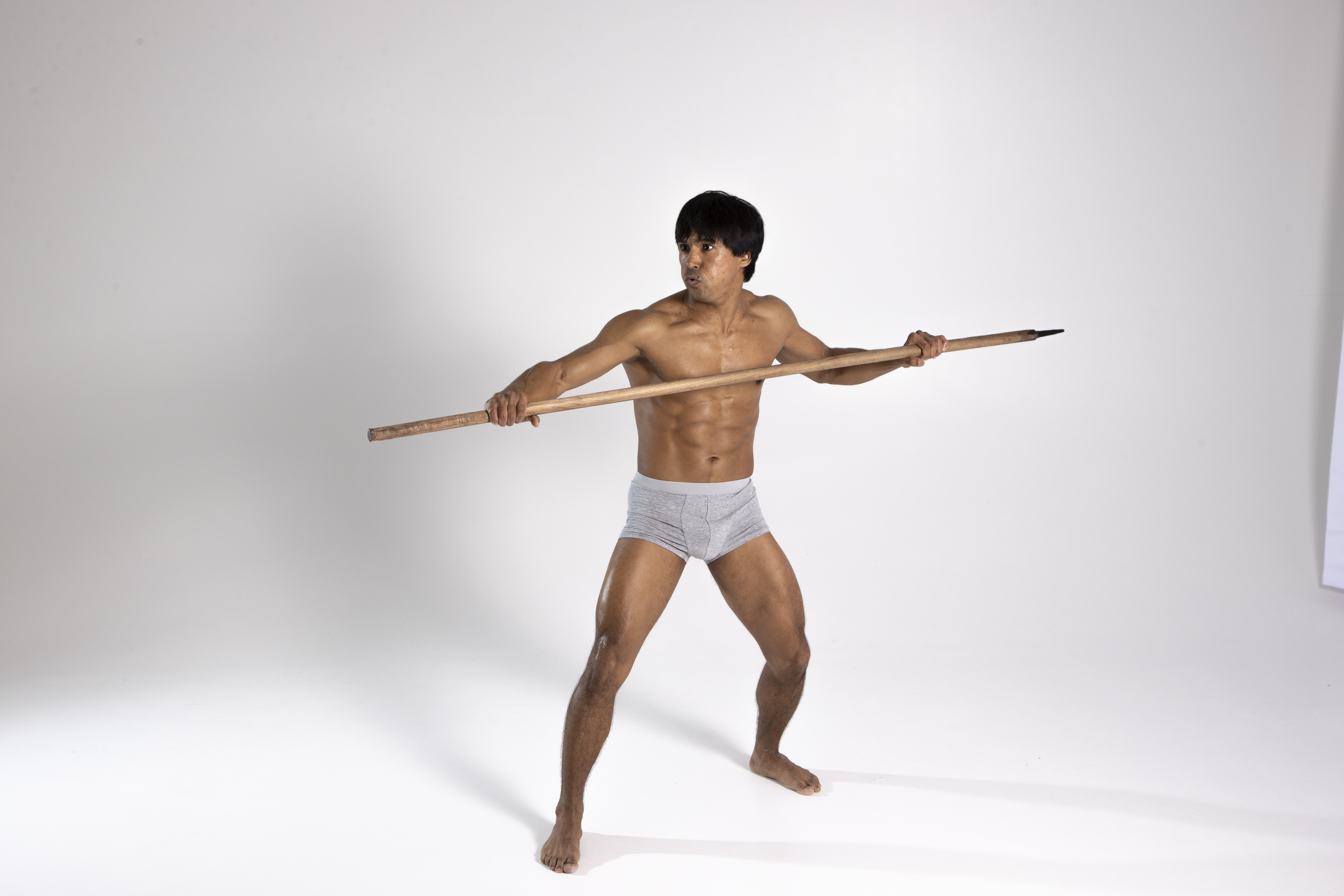 a shirtless man stands in a defensive stance holding a spear against a white background. he has dark hair and is wearing grey underwear.