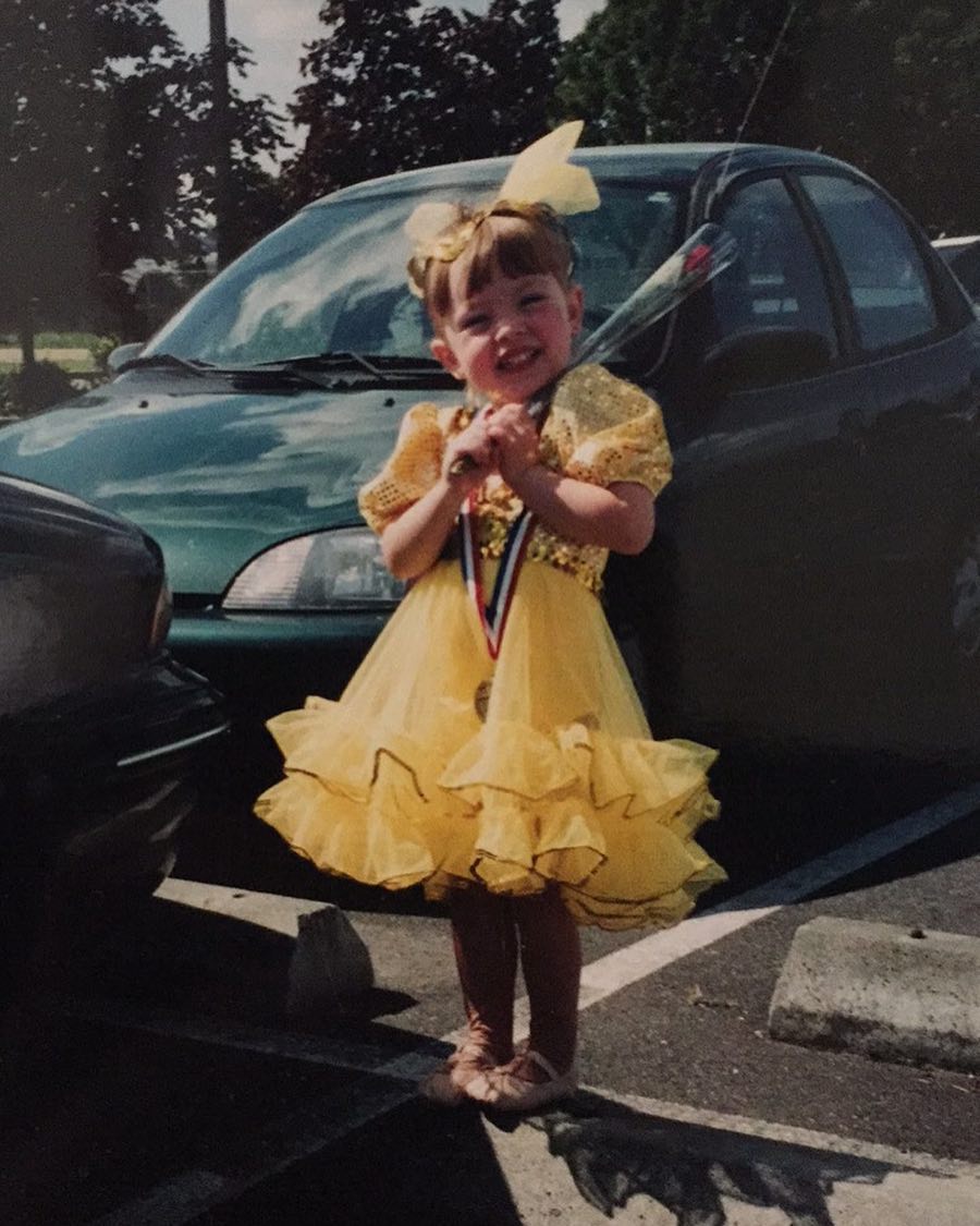 a photo of a young girl, possibly a toddler or preschooler, in a bright yellow ruffled dance dress. she is holding a single rose and a medal around her neck. she's standing in a parking lot in front of a darkcolored car, smiling happily.  the overall impression is one of a young girl having won a dance competition or recital. the setting suggests an outdoor event.