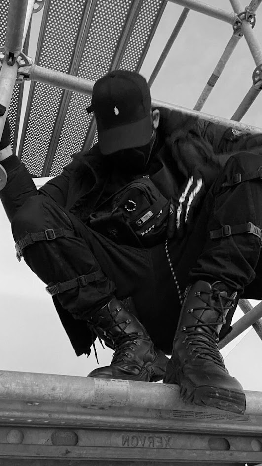 a black and white photo of a man in all black streetwear clothing. the man is wearing a black cap, black boots, and black pants. he is standing on a metal scaffolding structure, looking down at the camera.  the image is moody, and the man's style is edgy and cool.