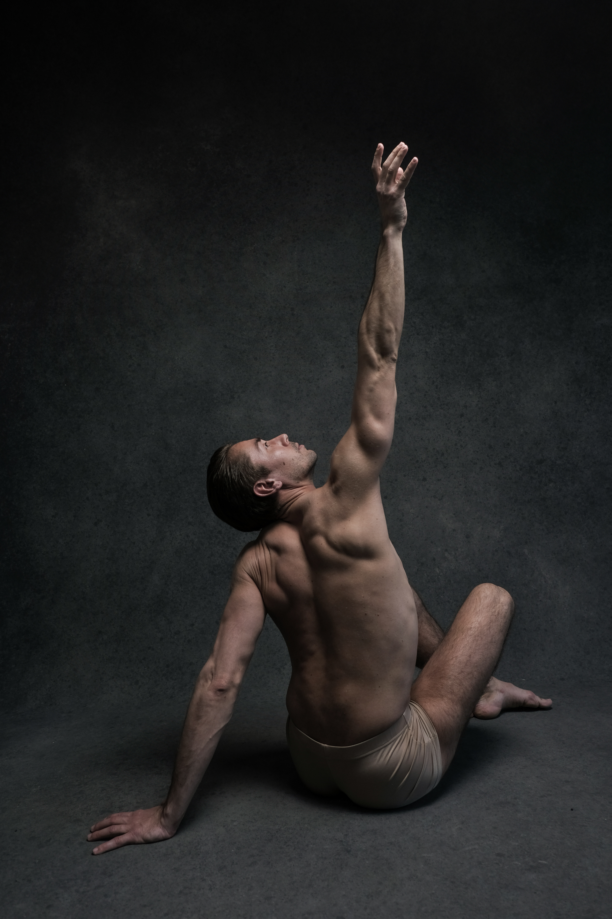 a muscular man, wearing only beige shorts, is captured middance pose against a dark studio background. his body is contorted with his left arm reaching upwards, his head tilted back, and his legs bent as if in a powerful leap or extension. the lighting is dramatic, highlighting the contours of his muscles and creating a sense of dynamic movement.