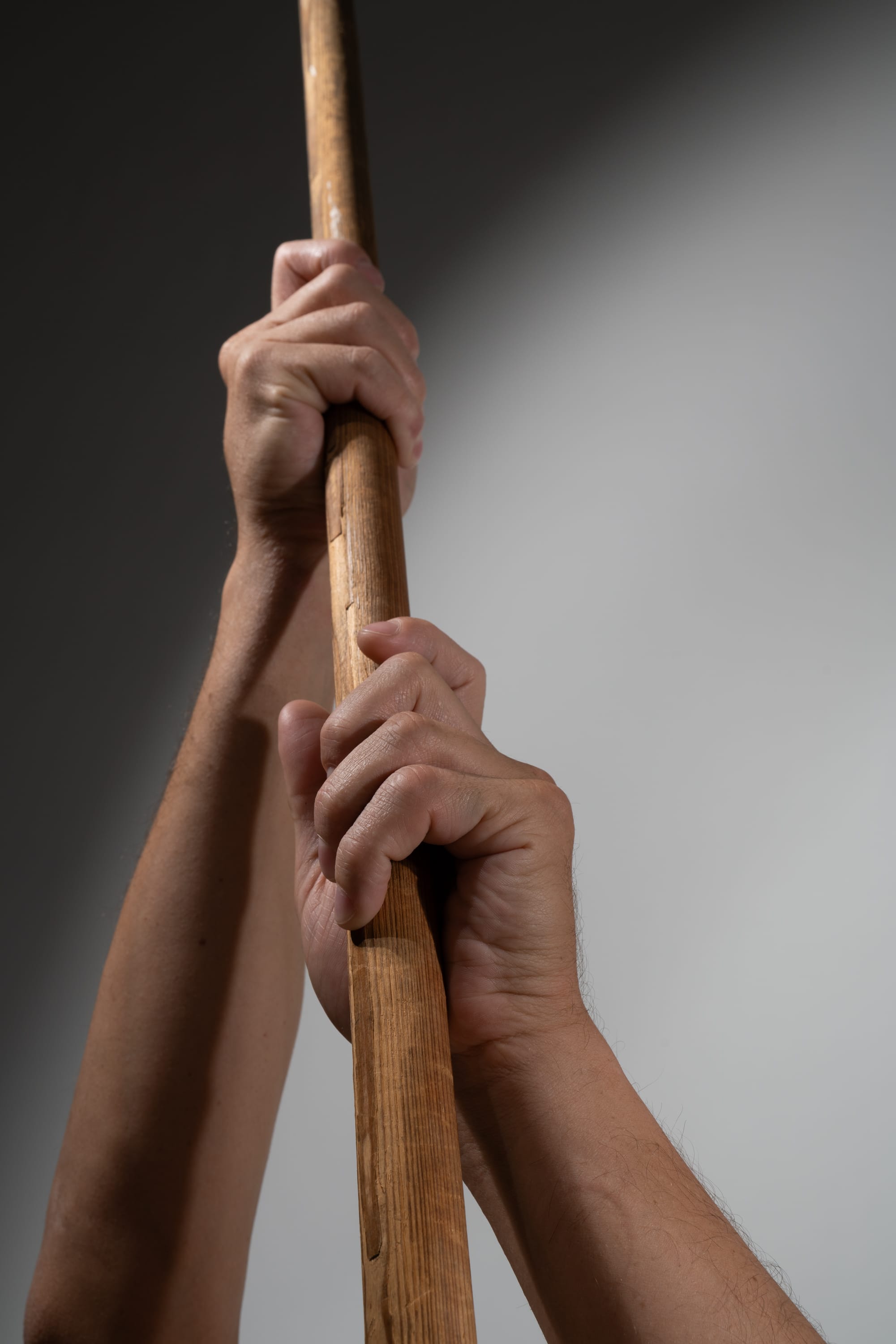 the image shows a pair of hands tightly gripping a long, brown, wooden pole against a gray and white background. the hands are reaching upwards, suggesting the pole is being held vertically. the lighting is bright, creating a studiolike effect.
