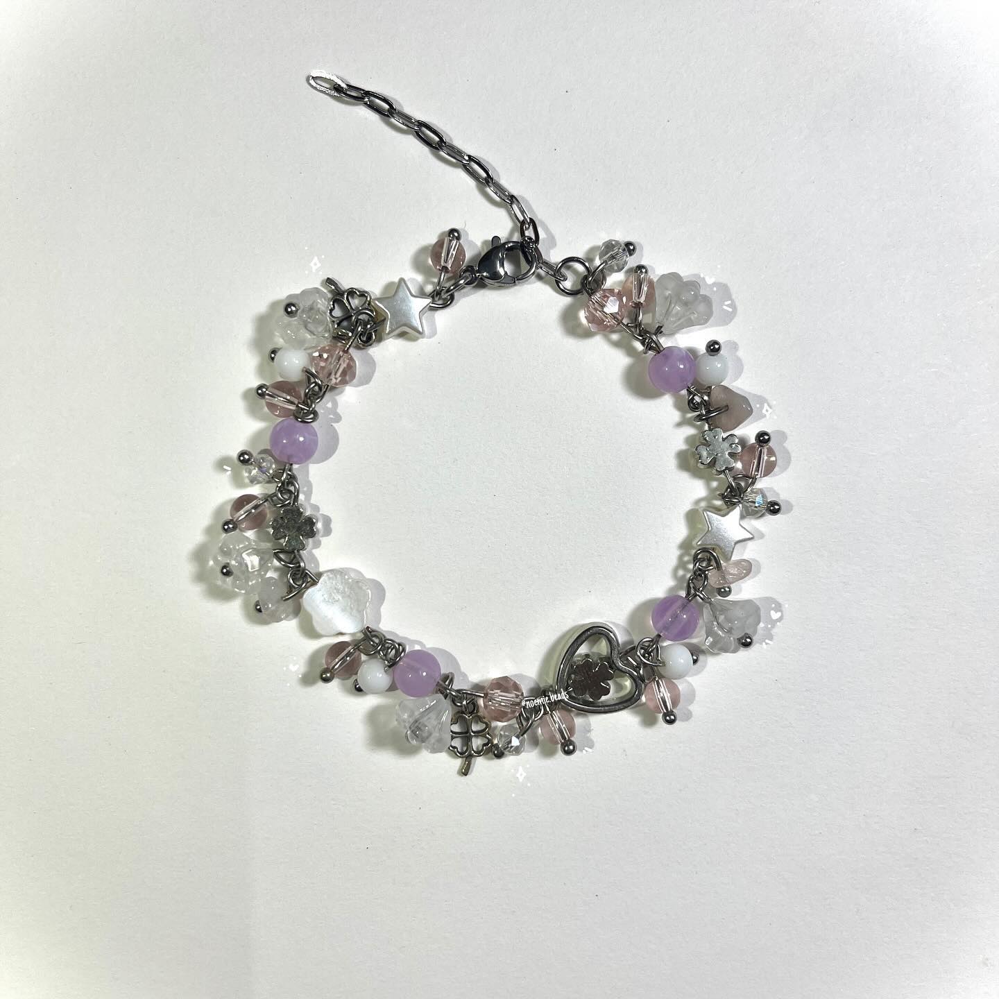 a handmade charm bracelet made with different beads and charms including purple, white, pink, and silver beads, a star charm, heart charm, and clover charm on a silver chain.