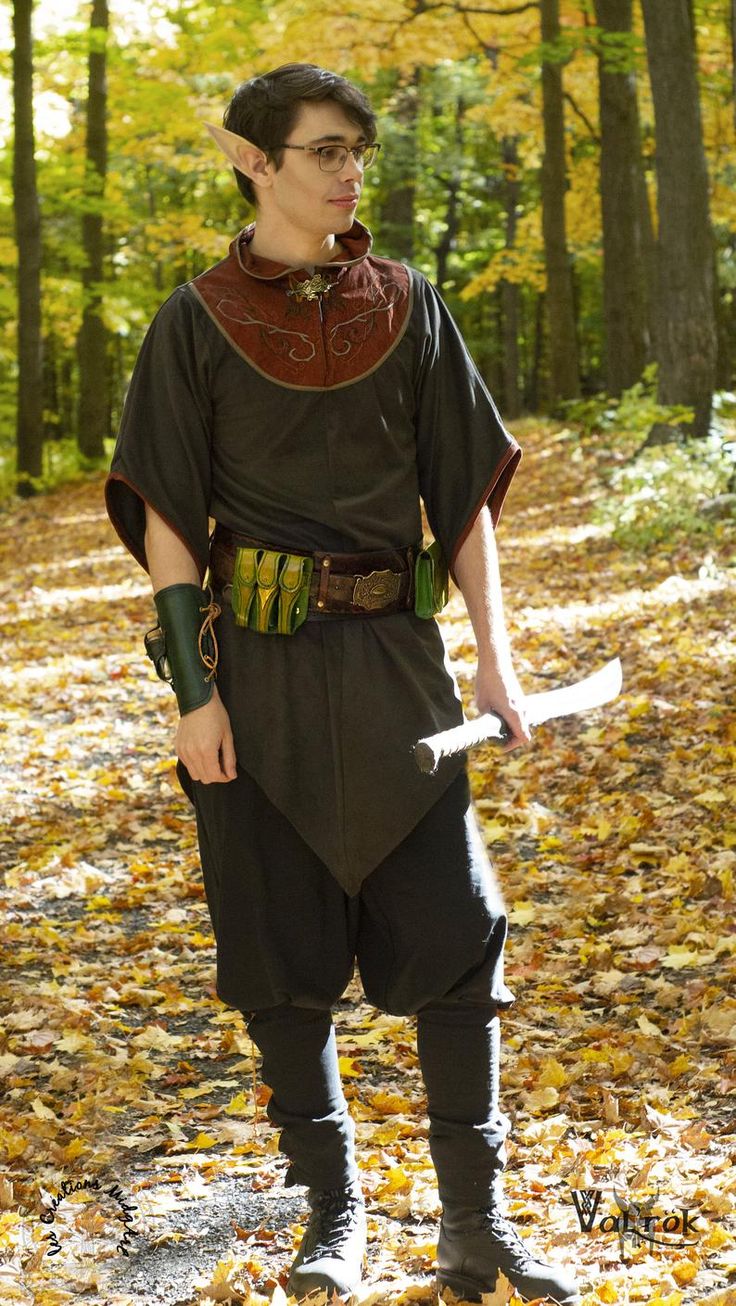 a fullbody shot of a male cosplayer dressed as an elf in a forest setting.  the elf is wearing a dark brown tunic with a reddishbrown embroidered collar, dark pants, and tall boots. he carries a short sword or dagger and wears several pouches on a leather belt. the forest is filled with autumn leaves, creating a vibrant backdrop. the elf looks to his right, slightly away from the camera. he has pointed ears and is wearing glasses.  the overall tone is one of fantasy and adventure.