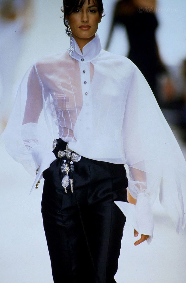 a woman is posing on a runway, wearing a highfashion outfit. she is wearing a white, sheer blouse with a high collar and buttondown front, paired with highwaisted black pants that feature decorative jewelry embellishments at the waist. she also has large, ornate earrings. the background is blurred, suggesting a fashion show setting.