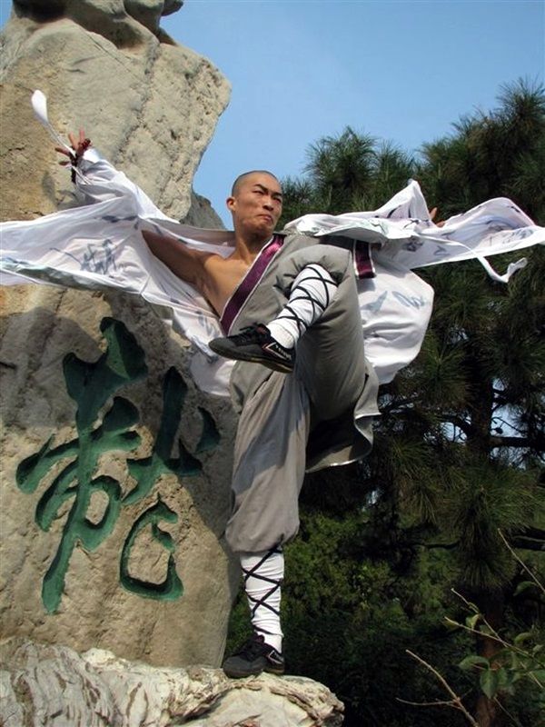 a young shaolin monk is demonstrating his kung fu skills. he is practicing a powerful leg kick in front of a large stone. the monk is wearing a white robe, and has his leg up high in the air, and his other leg is bent. his hand is out, ready to strike. in the background are mountains, trees, and foliage.