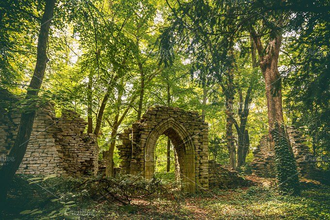 an old stone ruin stands amidst a lush forest. the ruins feature an arched doorway and crumbling walls, overtaken by nature. trees surround the structure, casting shadows and providing a backdrop of green foliage.