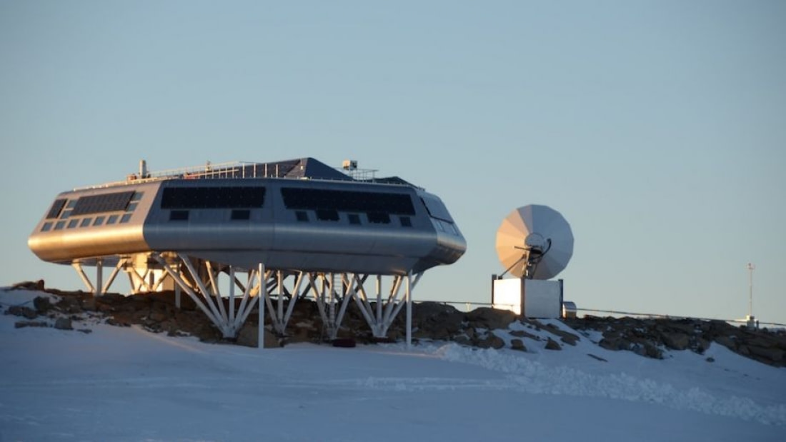 a satellite dish and a research station sit on a snowy mountainside. the station is a large, metal building with a flat roof and large windows. the satellite dish is white and round, and is pointed towards the sky. the sky is blue and there are snowcovered mountains in the background. the scene is one of desolation and isolation, but also of human ingenuity and determination.