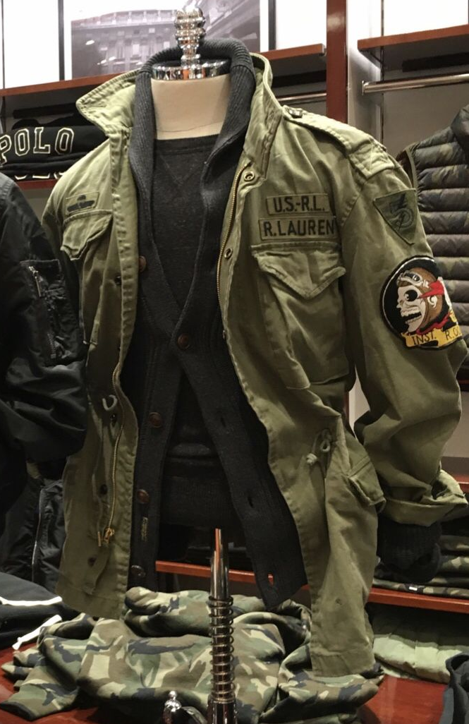 a closeup shot of a green military jacket on a mannequin in a clothing store. the jacket has a camo pattern on the sleeves and a patch that says "usrl r. lauren". it is displayed in a store setting with other clothing items in the background. the jacket is hanging on a mannequin with a black shirt underneath. the photo is taken from a low angle, and the focus is on the jacket. the image is slightly blurred.