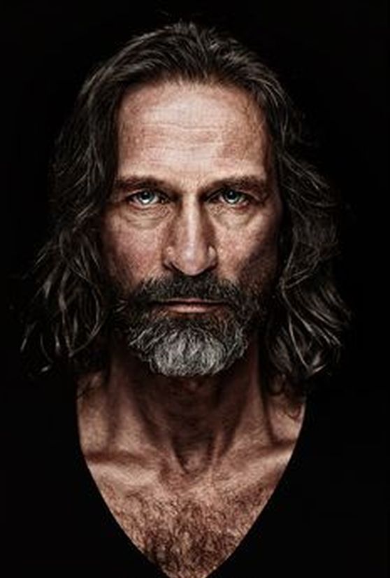 a studio portrait of a seriouslooking mature man with a beard and long hair. he is wearing a black shirt and has chest hair visible. the background is dark.