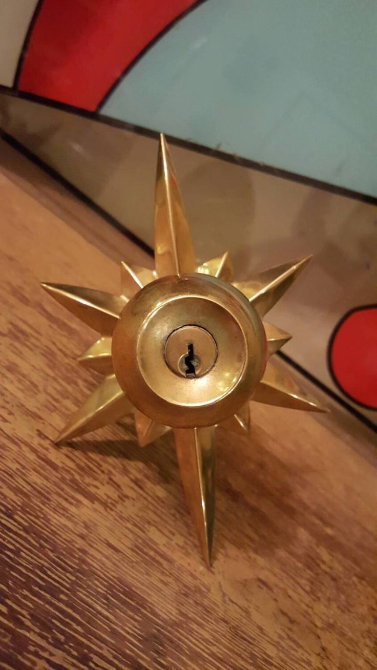 a gold, star shaped door knob with a circular keyhole in the center. it appears to be a vintage brass door handle.  the door knob is sitting on a wood surface and the background is blurry and multicolored.