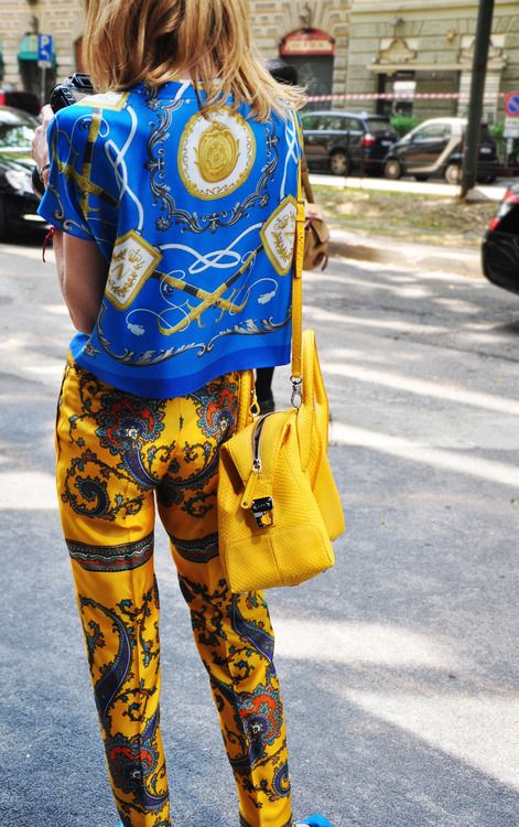 a woman is walking down a city street. she is wearing a vibrant blue silk shirt with a paisley print and matching yellow paisley print pants.  she carries a bright yellow handbag slung over her shoulder. the photo is taken from behind, showing her back.  the overall style is a bold and fashionable summer look.