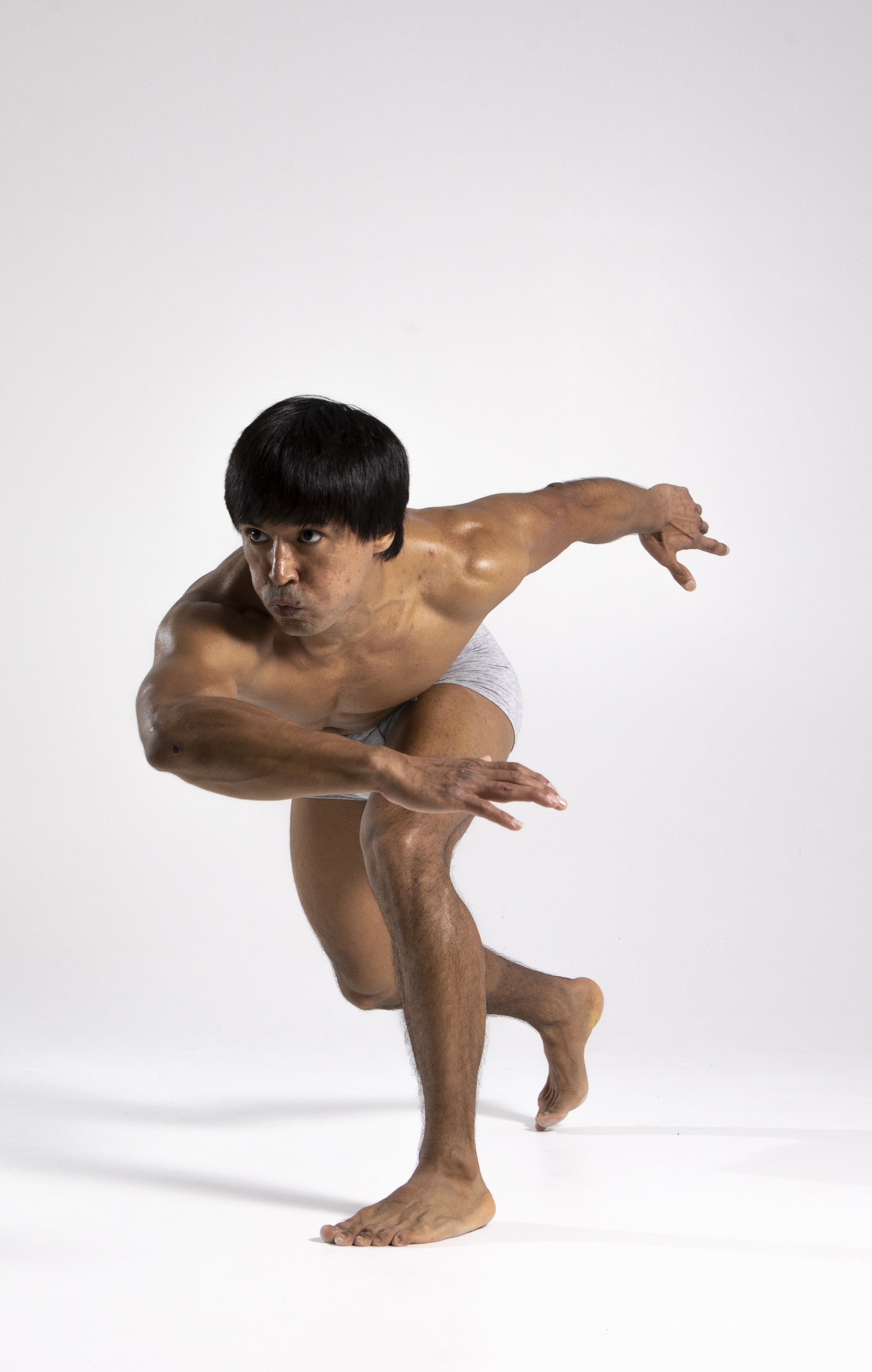 a muscular, barechested asian man is captured in a dynamic action pose against a white background. he is in a low, runninglike stance, with one leg forward and bent, the other trailing behind. his arms are extended in front of him, hands open as if bracing for impact. he has short, dark hair with bangs. the image emphasizes his physique and the energy of the movement.