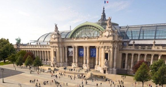 the image showcases the grand palais in paris, france. this iconic beauxarts style building is characterized by its massive glassroofed central nave and its flanking symmetrical wings.  the photo is taken from a slightly elevated viewpoint, offering a broad perspective of the building's facade and the surrounding plaza. many people are visible strolling around the grand palais, adding a sense of scale and daily life to the scene. the bright, sunny day highlights the architecture's intricate details and the clear sky enhances the overall aesthetic appeal. the overall impression is one of grandeur, history, and the lively atmosphere of a parisian public space.
