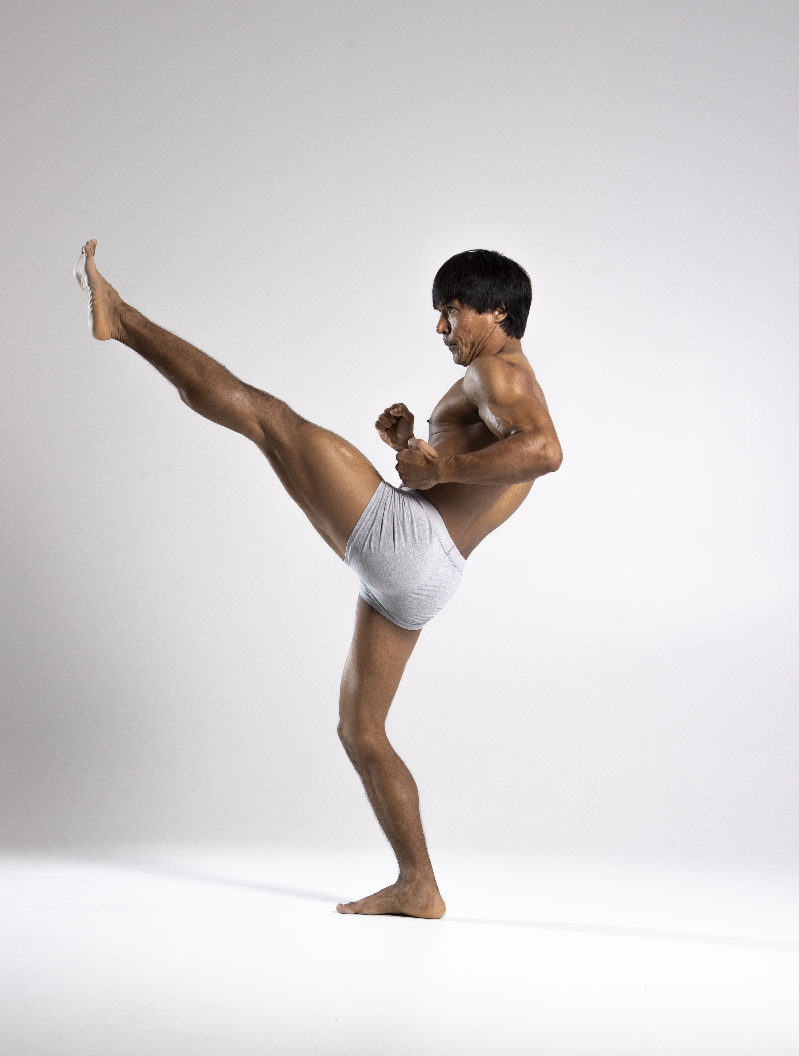 a shirtless man in lightcolored underwear is captured in a studio shot performing a high kick. he is barefoot, and his body is angled as he executes the martial arts move. his expression shows focus and intensity. the background is plain and neutral, emphasizing the figure and the action.
