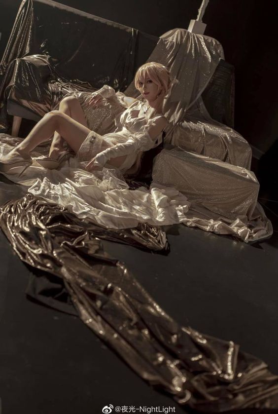 woman in a white, flowing gown lies on a white chaise lounge and the floor. she is surrounded by shimmering gold fabric that drapes over the furniture and cascades across the floor.  the setting is dark, illuminated by warm, artificial light that creates dramatic highlights on the fabric and the woman's form. her pose is languid and elegant.