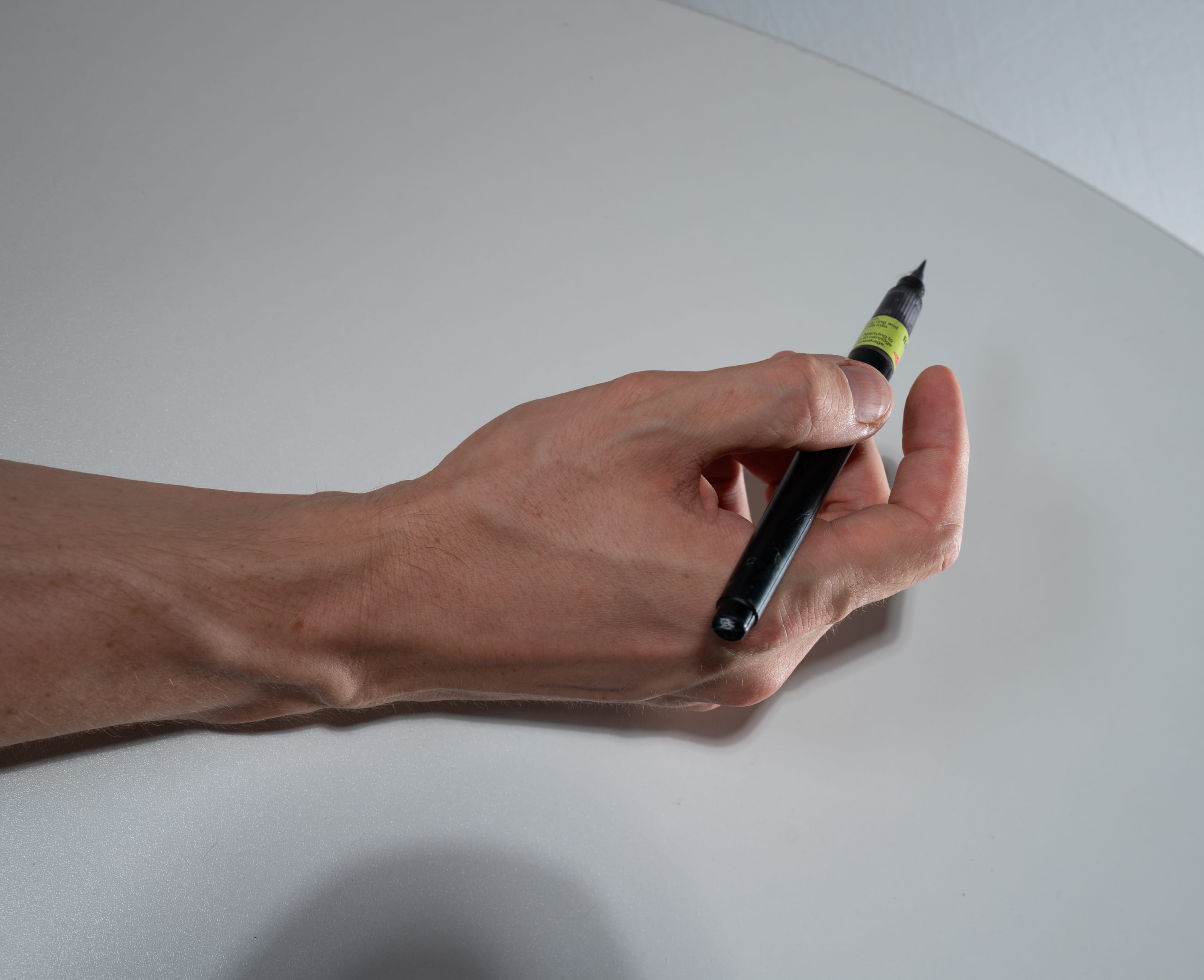 the image shows a caucasian hand holding a black pen on a white surface. the pen has a yellow sticker label on it. the lighting is artificial, suggesting a studio setting. the closeup perspective emphasizes the details of the hand and the pen.
