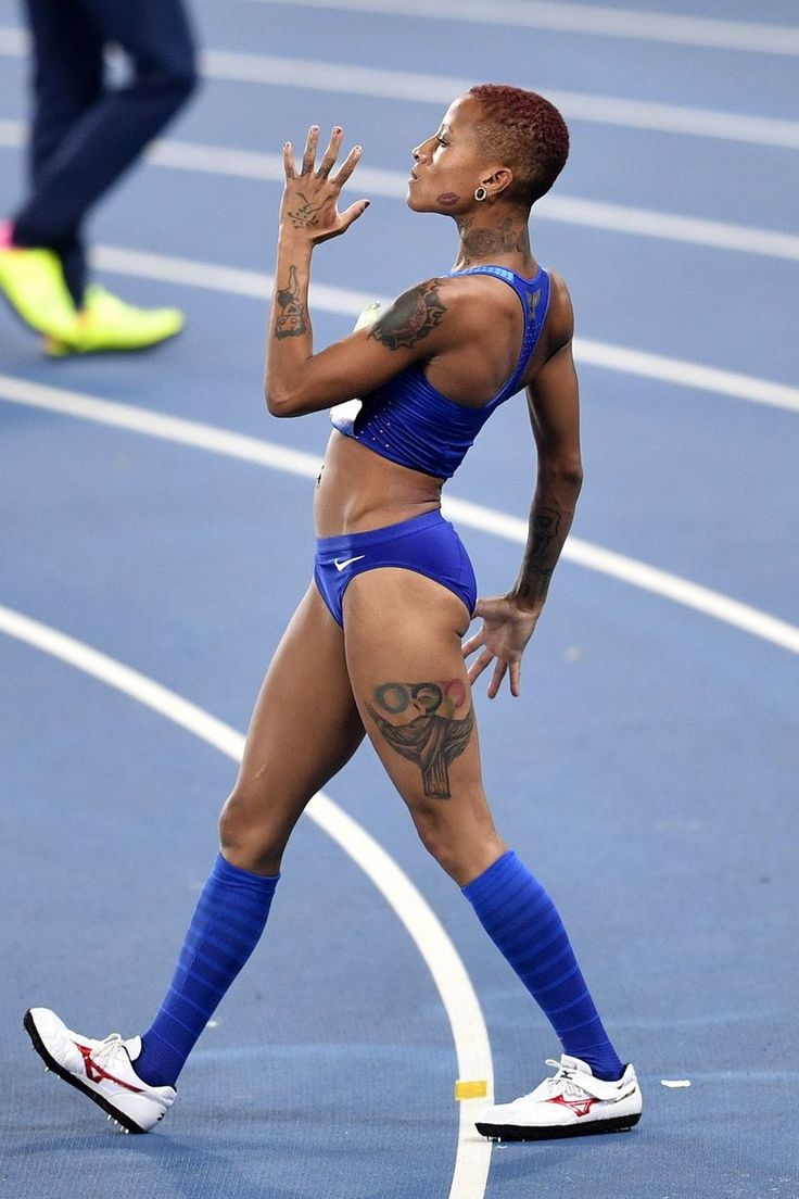 a female track and field athlete in a blue uniform and socks celebrates her victory on the track.  she is looking upward, with one arm raised, and has visible tattoos on her arms and legs. her expression is one of triumph and exhilaration.  the background is a blue running track. the overall impression is one of athleticism, skill, and achievement.