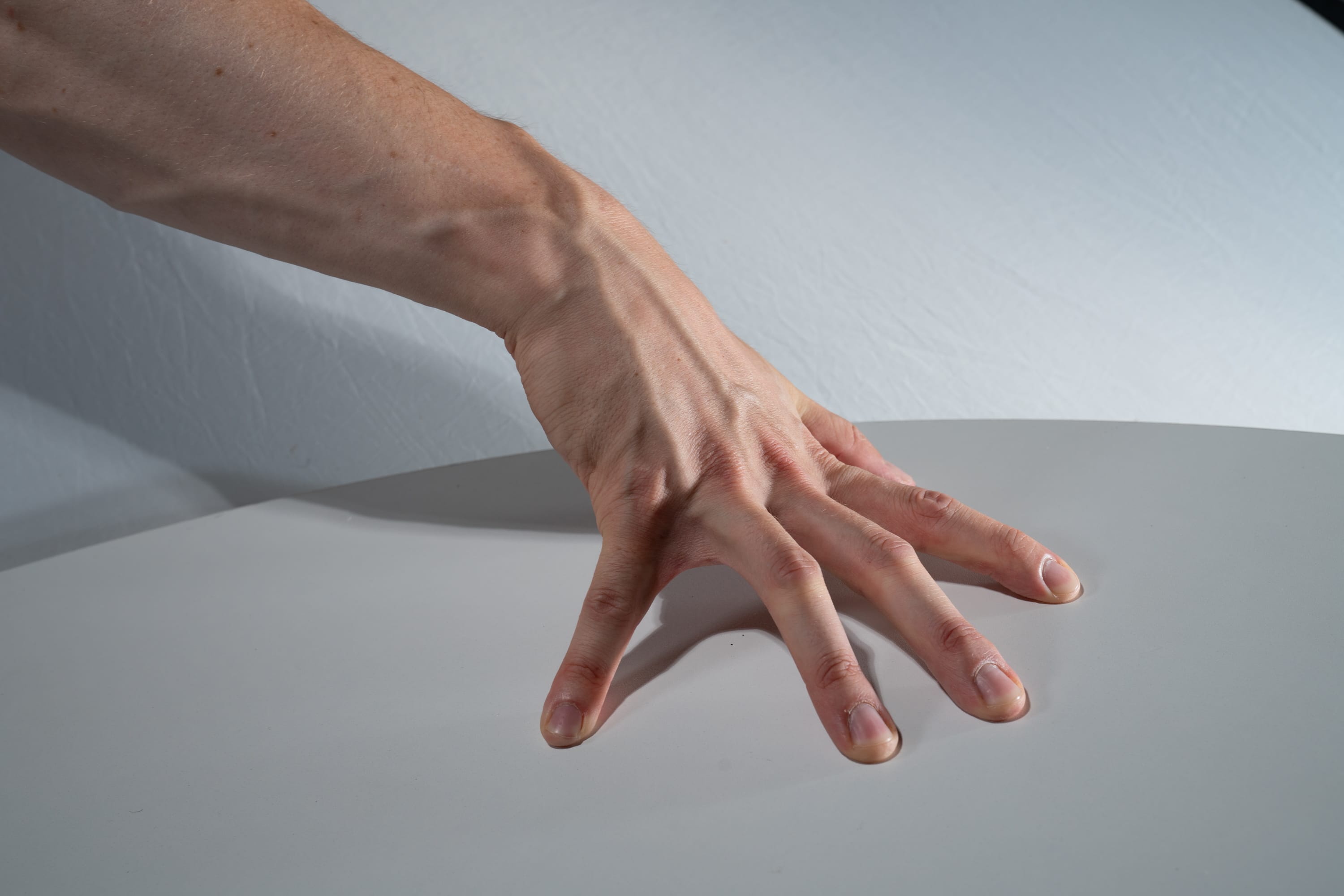 a man's hand is spread out on a white table, illuminated by studio lighting. the closeup perspective highlights the details of his skin and veins.