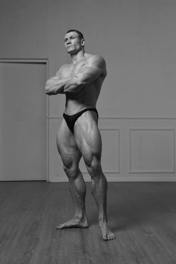 a black and white full shot of a muscular male bodybuilder standing in a studio. he is posing with his arms crossed, showcasing his welldefined physique. he is wearing a darkcolored brief. the background is a simple, lightcolored wall with white molding. the floor is wood. the overall mood is serious and confident. the image is reminiscent of classic bodybuilding photography.