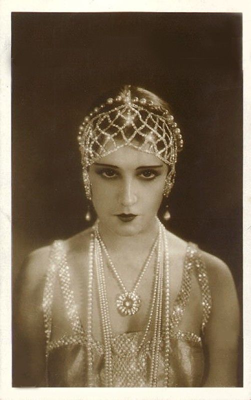a vintage portrait of a woman wearing a flapper outfit and pearls. the woman has on a headband, a pearl necklace, and a beaded dress. her look is vintage and glamorous. the background is a simple black studio background. the image is a historical snapshot of 1920s fashion.