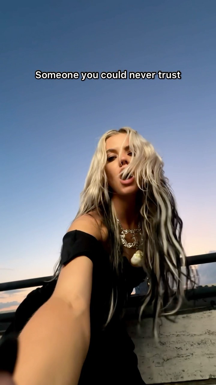 a lowangle shot of a woman with blonde hair and black streaks wearing a black dress and jewelry, taking a selfie against a sky backdrop. the text "someone you could never trust" is overlaid on the image.