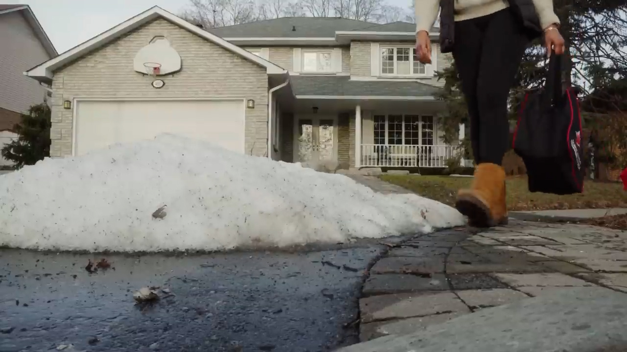 a lowangle view of a person walking on a paved driveway, past a large pile of melting snow. the snow is piled high, almost reaching the top of the driveway. the person is wearing black pants, brown boots, and carrying a black and red bag. the background is a twostory house with a basketball hoop on the garage door. the sky is overcast, and the overall lighting is dim.