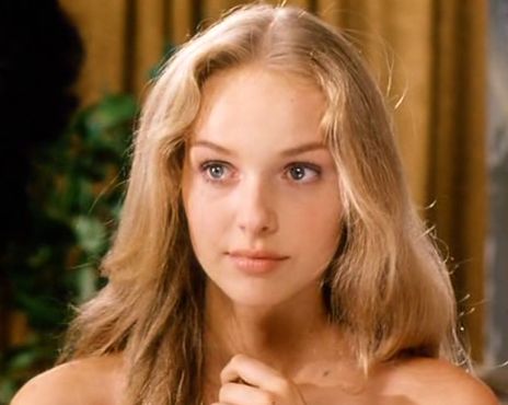 a portrait of a fairskinned woman with blonde hair and blue eyes. she has a gentle expression. she is depicted from the shoulders up. her hair is long and wavy. she has fair skin. her shoulders are bare. her hand is visible near her chest. the background includes green foliage on the left and a curtain on the right. the scene appears to be indoors.