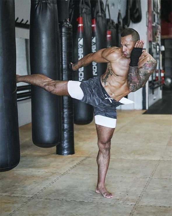 a man in a gym, wearing shorts, is working out by kicking a punching bag.  he has a powerful build and a muscular physique. the man is training in martial arts.