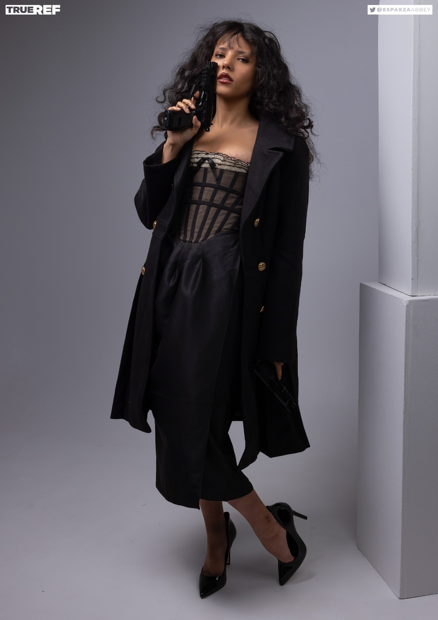 a full shot of a woman in a black coat and dress, holding a gun to her head. she has dark hair and is wearing black heels. she is posing in a studio setting on a gray background. the overall look is stylish, elegant, and powerful.