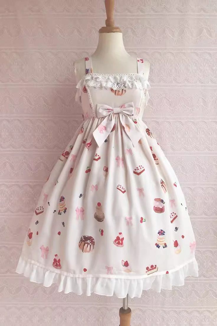 this is an image of a lolita fashion dress. the dress is white with a pink and red dessert print. it has a square neckline with lace trim, a bow at the waist, and a ruffled hem. the dress is shown on a mannequin in front of a pink wall.