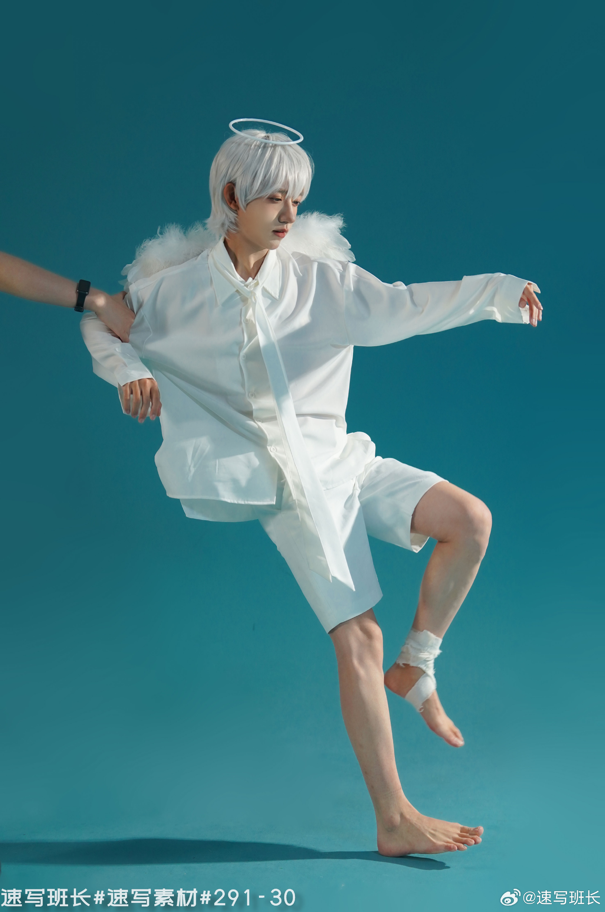 an asian male with silver hair is dressed in a white angel costume, complete with a halo and feather wings, posing against a light blue background. he is barefoot and wears a bandage on his right ankle. the lighting is bright and artificial, creating a studiolike setting. the image captures him midpose with his arms outstretched, as if in motion. a hand reaches out from the left side of the frame, gently touching his arm. the overall impression is one of playful theatricality and fantasy.