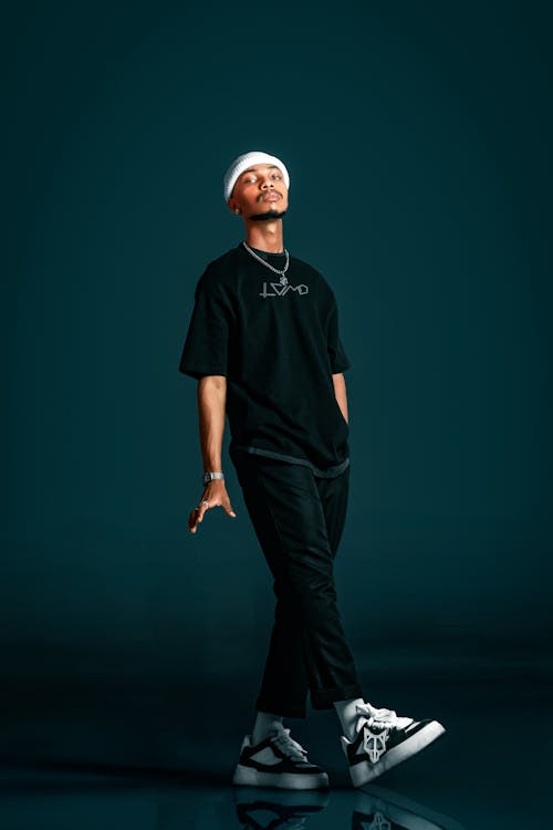 the image showcases a young africanamerican male model dressed in stylish streetwear. he is wearing a black tshirt with a subtle logo, black pants, white beanie, and white and black sneakers.  he’s also wearing a silver necklace.  he’s posed in a relaxed yet confident manner against a dark teal backdrop. the overall aesthetic is trendy and urban. the lighting highlights his features and the details of his outfit.