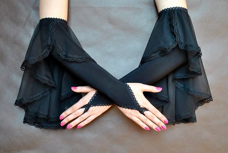 the image shows a pair of crossed hands wearing black fingerless gloves with lace and mesh detailing, against a gray background. the person has pink nail polish.