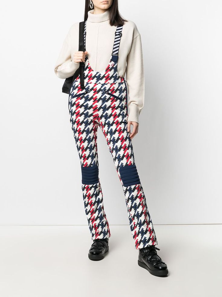a woman model wearing a white, red, and blue patterned ski suit with black boots. she is standing in a white background and the outfit is a classic ski wear.