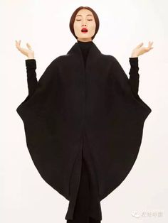 a woman in a black dress poses dramatically with arms outstretched. the dress features a long flowing cape with a deep vneck. she wears a bold red lipstick and looks straight into the camera.