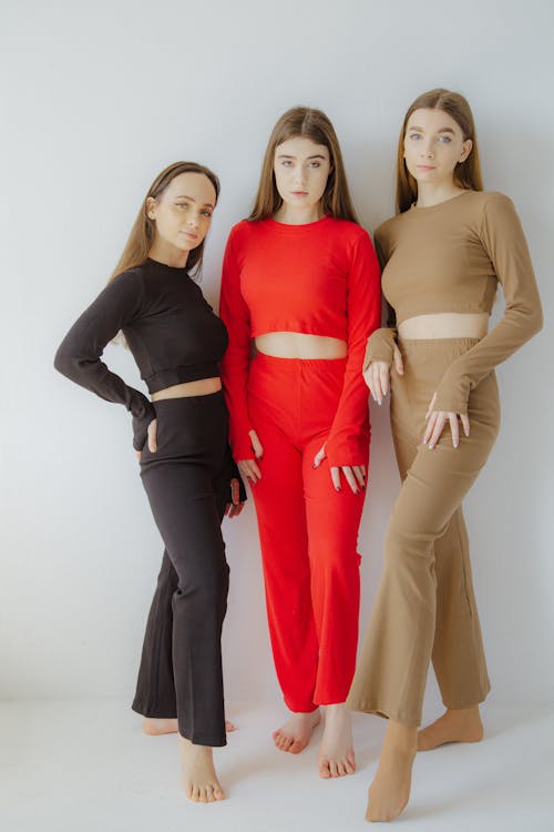 three women wearing matching loungewear sets in different colors stand barefoot in front of a white wall. the woman on the left wears a black crop top and pants, the woman in the middle wears a red set, and the woman on the right wears a brown set. all of the sets consist of a longsleeved crop top and wideleg pants. the women are facing forward and looking at the camera.