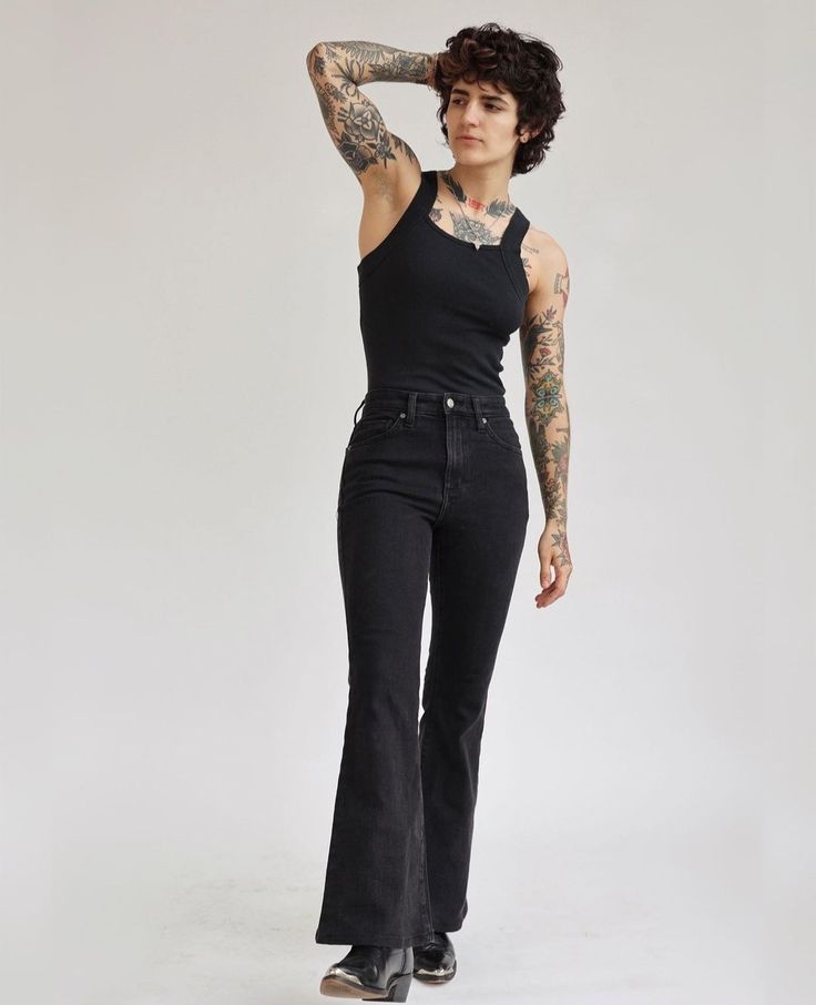a fullbody shot of a young woman with extensive tattoos modeling a black ribbed tank top and dark wash bellbottom jeans. she has short, curly brown hair and is posing against a plain white background.  she's wearing what appear to be westernstyle ankle boots and has one hand resting behind her head. the overall style is casual yet fashionable.