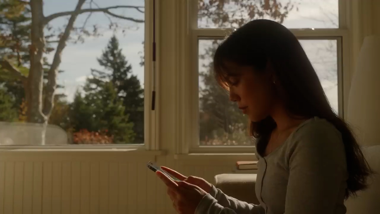 a young woman sits by a sunlit window, engrossed in her phone. warm, natural light bathes the scene, highlighting the autumnal colors of the trees visible outside. the image evokes a sense of quiet contemplation and connection.