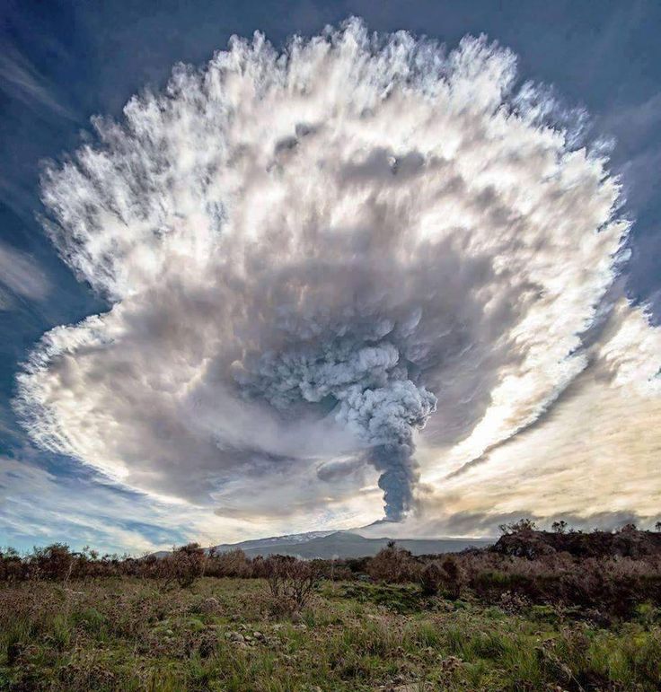 a massive ash plume billows out of the fagradalsfjall volcano in iceland, creating a dramatic and aweinspiring sight against a backdrop of blue sky and fluffy white clouds. the plume stretches high into the air, a testament to the power of nature. the surrounding landscape is blanketed in green vegetation, creating a striking contrast to the dark, ominous cloud. the image captures the raw power and beauty of a volcanic eruption.
