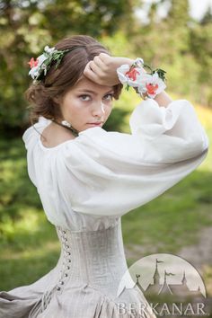 a young woman with flowers in her hair, dressed in a flowing white medievalstyle blouse and corset.  she's posed outdoors in a soft, natural light that highlights the delicate fabric of her outfit and the flowers adorning her hair. the image has a romantic, ethereal quality.