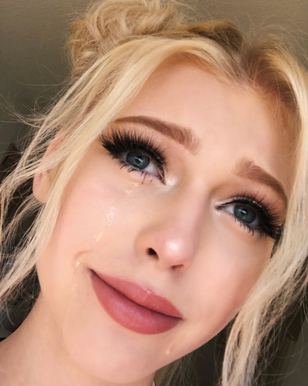 a closeup selfie of a young blonde woman with blue eyes, wearing heavy makeup and long eyelashes. she appears to be crying, with tears visible on her face. her expression suggests sadness or emotional distress.