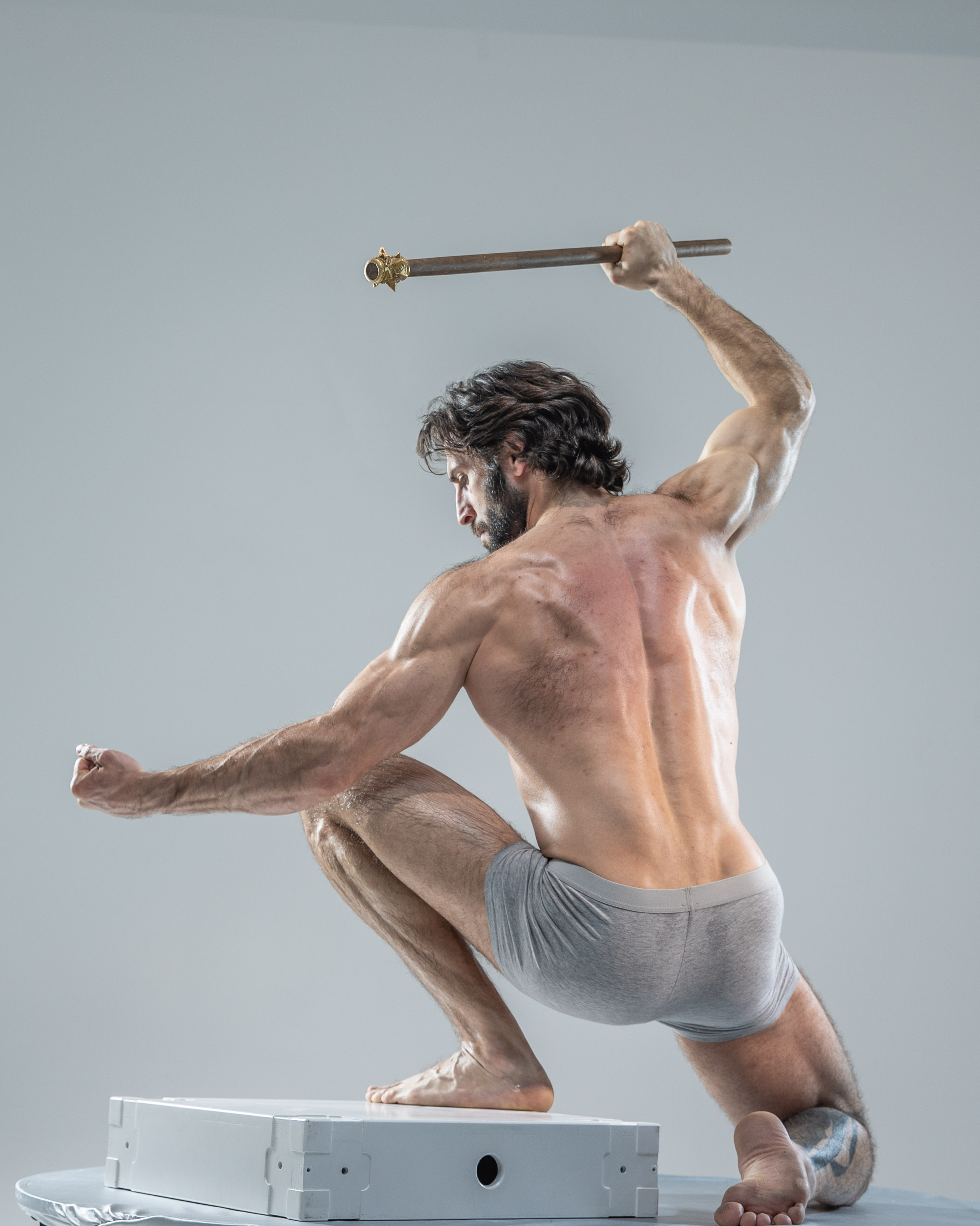 he image showcases a muscular man in a dynamic action pose, captured against a neutral gray studio backdrop. he is positioned with his back to the camera, his body arched in a display of strength and agility. the man holds a wooden staff, suggesting a warriorlike persona. the lighting is bright and soft, highlighting his welldefined musculature and the tension in his pose. the overall image evokes a sense of power, movement, and determination.