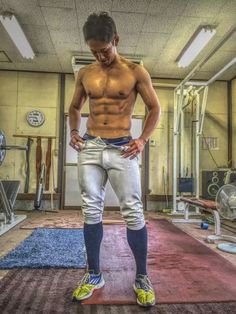 a muscular, shirtless man stands in a gym setting. he's wearing white baseball pants and black compression socks and shoes. the gym appears to be small and cluttered with various workout equipment.  he appears to have just finished or is about to begin a workout routine.