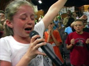 a young girl is singing into a microphone at a religious event. she appears to be crying with emotion. in the background, there is an audience of people watching.