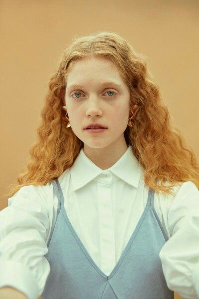 closeup portrait of a young woman with long, curly ginger hair. she is wearing a white collared shirt layered over a light blue sleeveless dress. her expression is calm and serene. the background is a soft beige.  she is wearing gold earrings. the overall style is minimal and elegant.