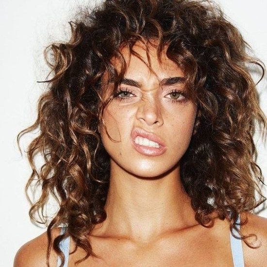 a young woman with curly brown hair and clear skin makes a funny face for the camera. she has freckles, brown eyes and is wearing makeup. she is wearing a blue bra. she is in a studio setting with a white background.