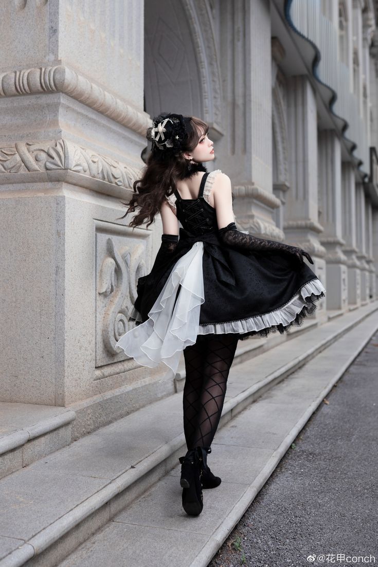 a woman wearing a black and white dress, black stockings and boots is walking on a pavement next to a building. she is looking up and away from the camera. she is wearing a black headpiece. the building is made of stone and has arches.