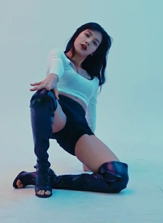 a woman wearing high black boots, black shorts, and a white top poses on a studio floor. the lighting is blue and the background is white.